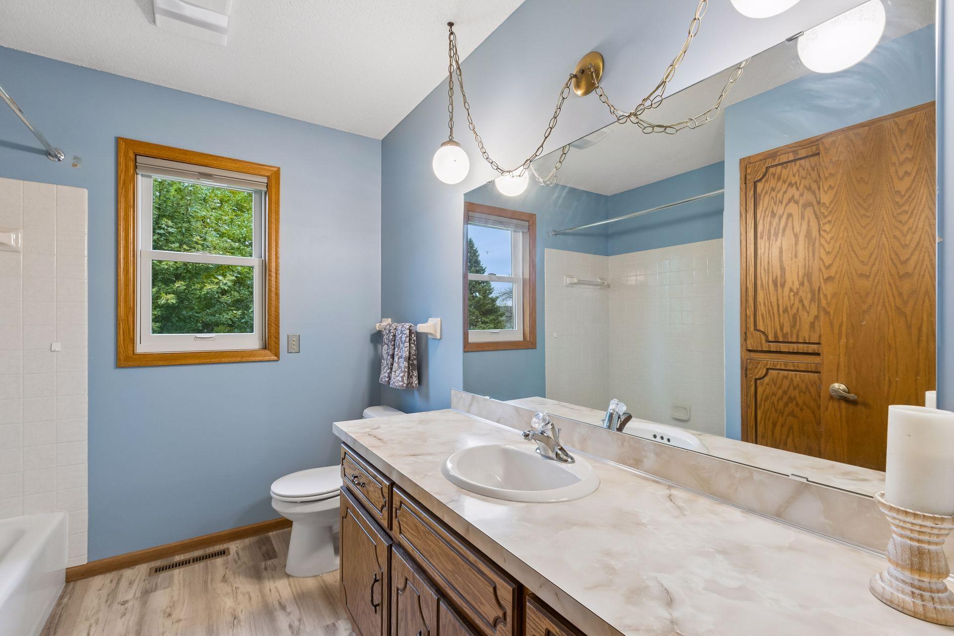 Bathroom with blue walls, a long wood vanity, marble-look counter, wide mirror, and tiled tub and shower.