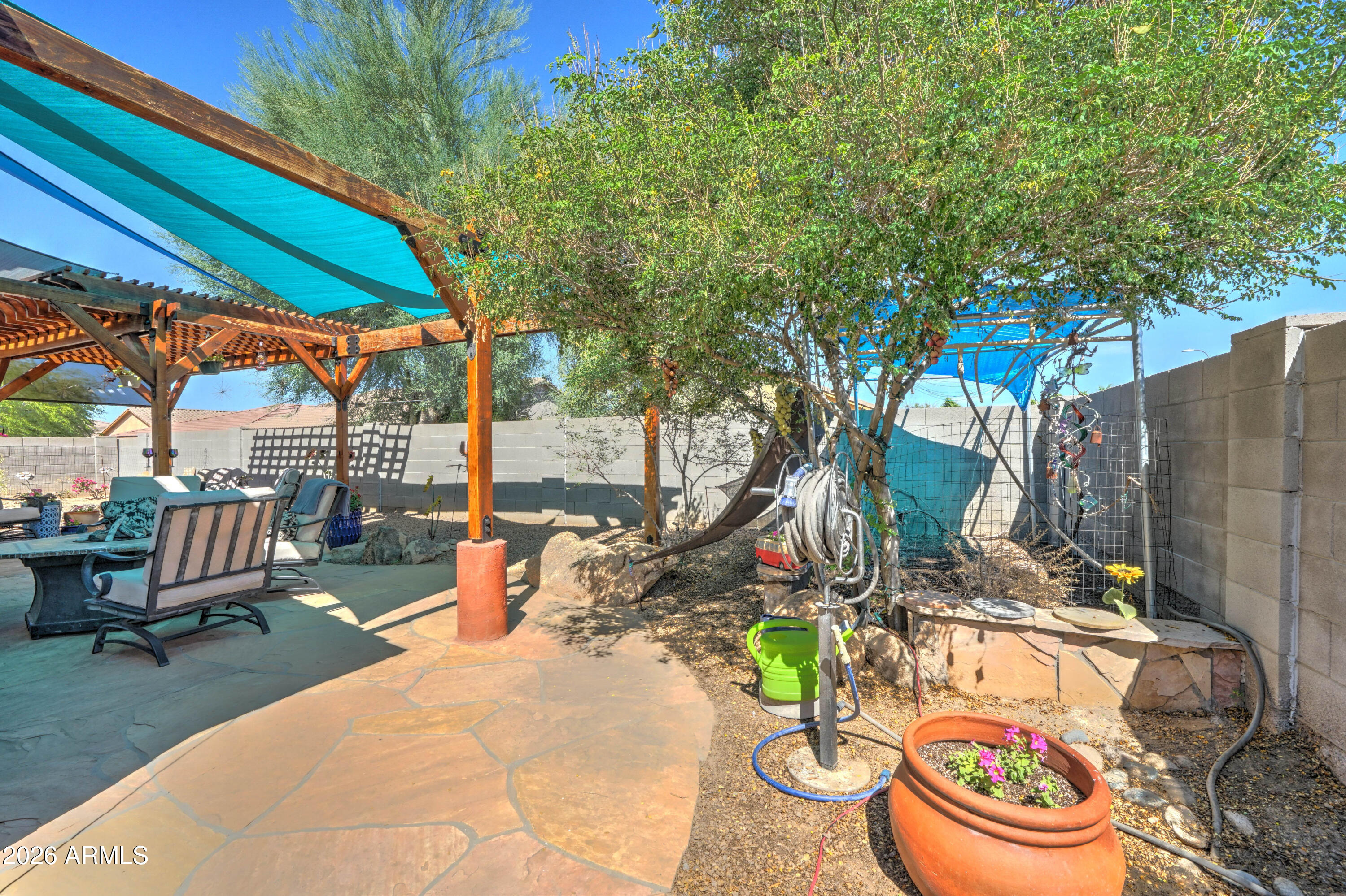Backyard patio with pergola, turquoise shade cloth, outdoor seating, and a leafy tree.