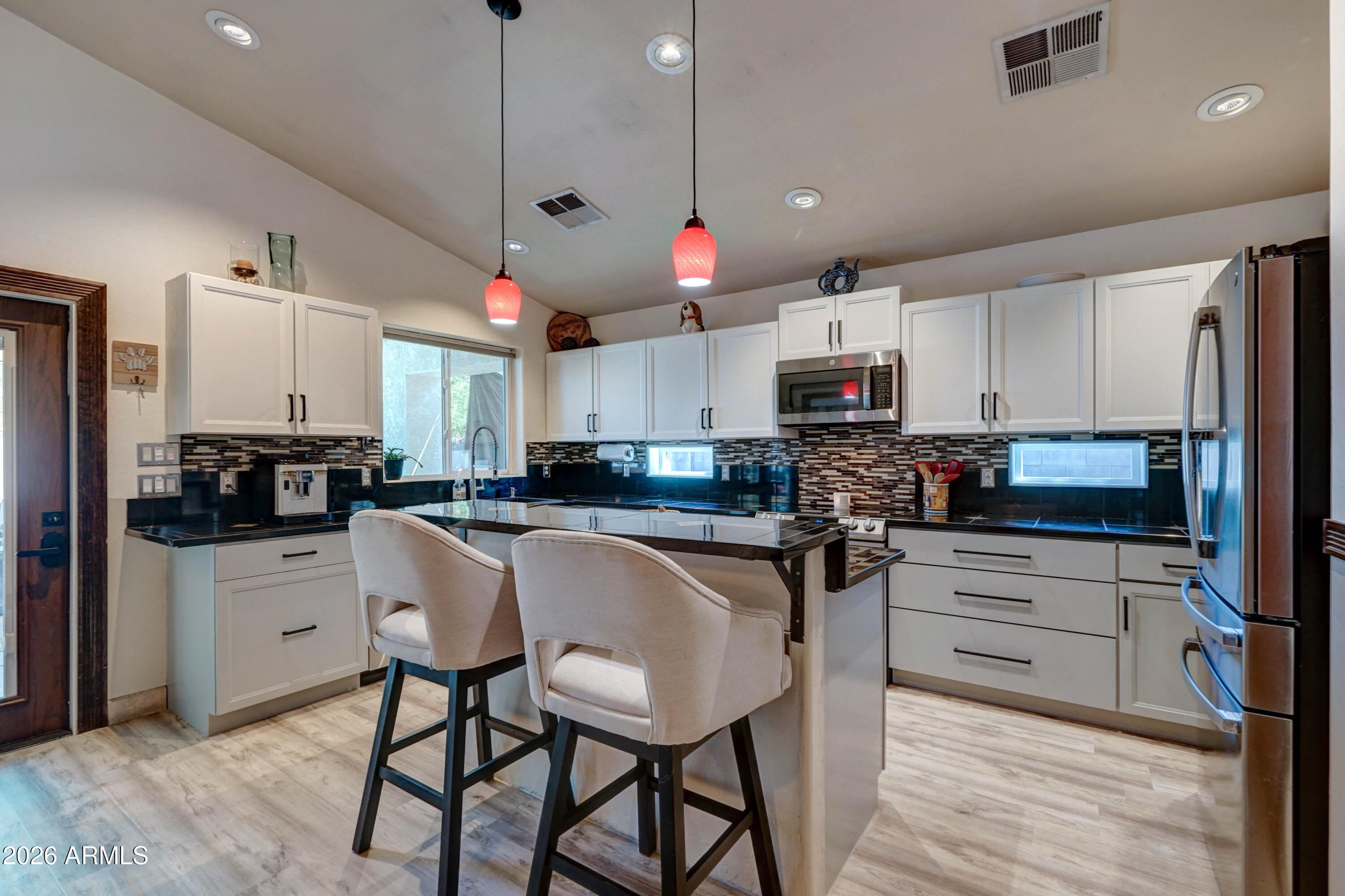 Kitchen with a central island, two upholstered barstools, white cabinets, and red pendant lights.