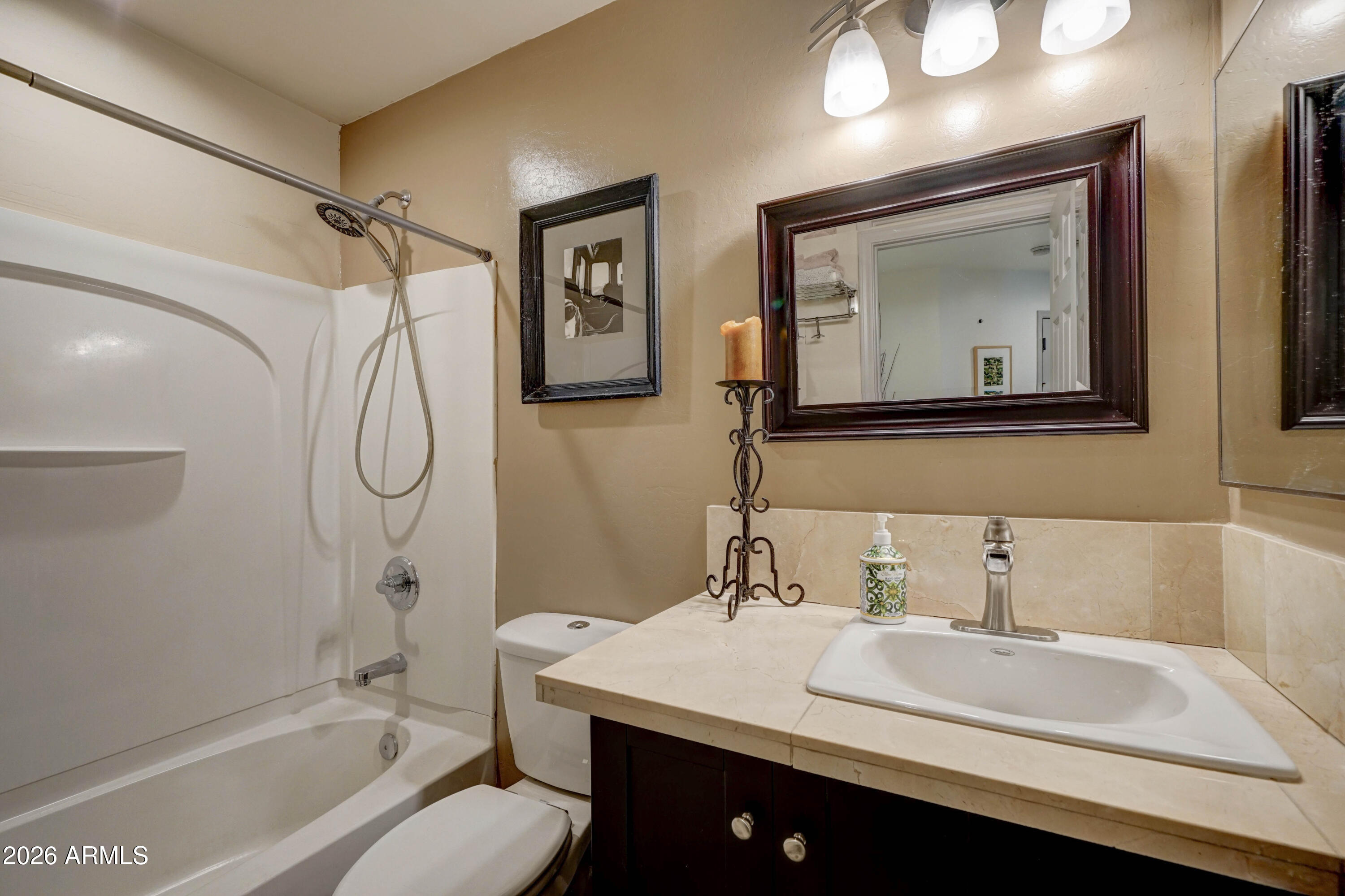 Bathroom with a white tub and shower beside a dark wood vanity with a cream stone countertop.