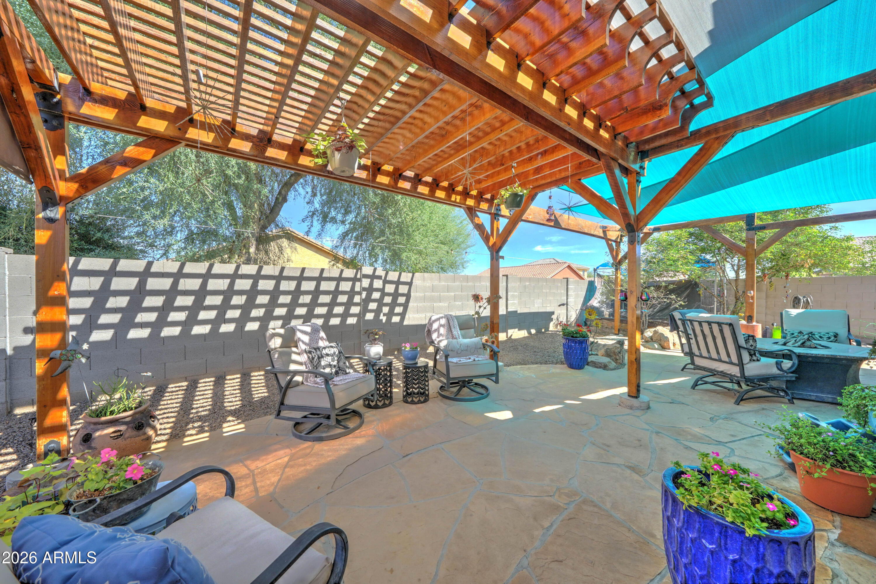 Backyard patio beneath a wooden pergola, furnished with lounge chairs and colorful potted flowers.