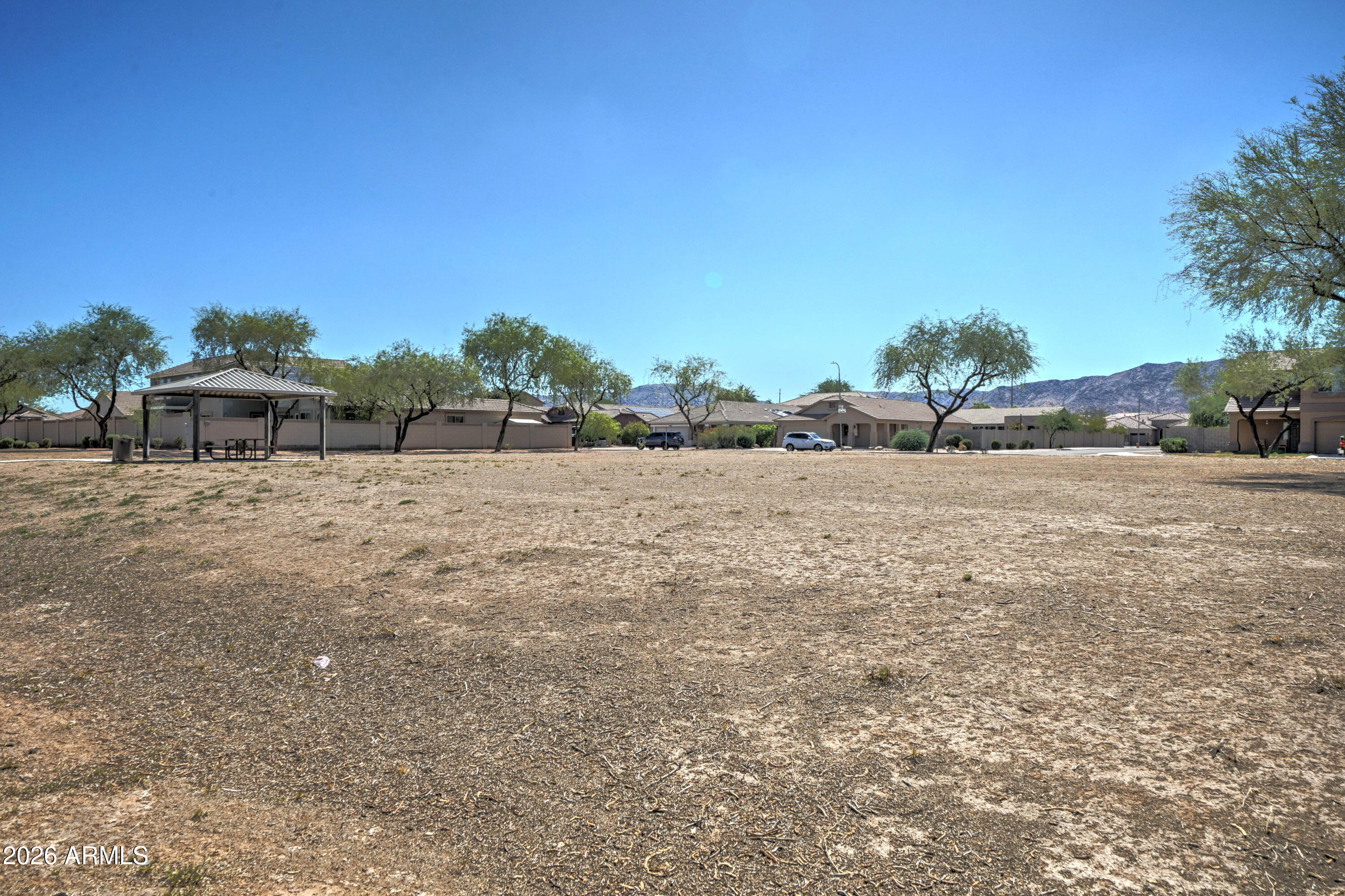 Dry open field with a covered picnic shelter, homes, and desert mountains in the distance.