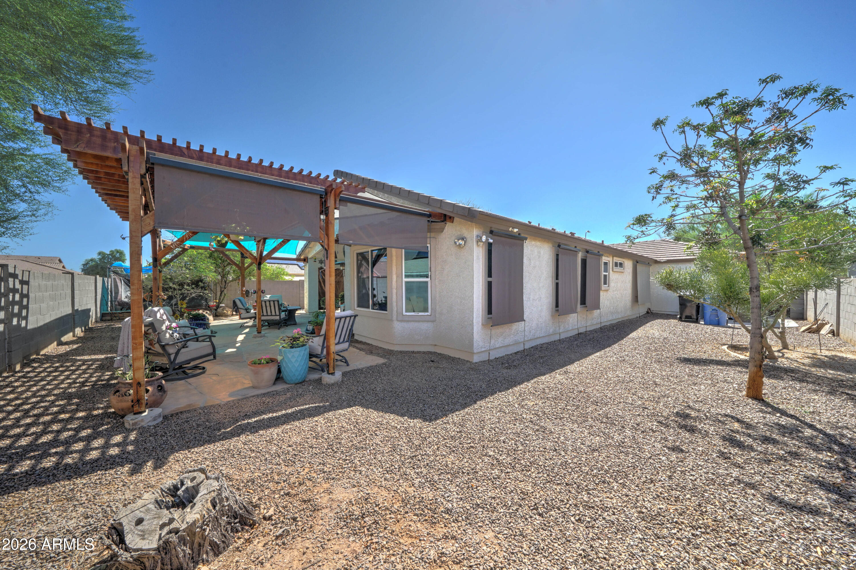 Home’s rear exterior with a shaded pergola patio, seating, gravel yard, and mature tree.