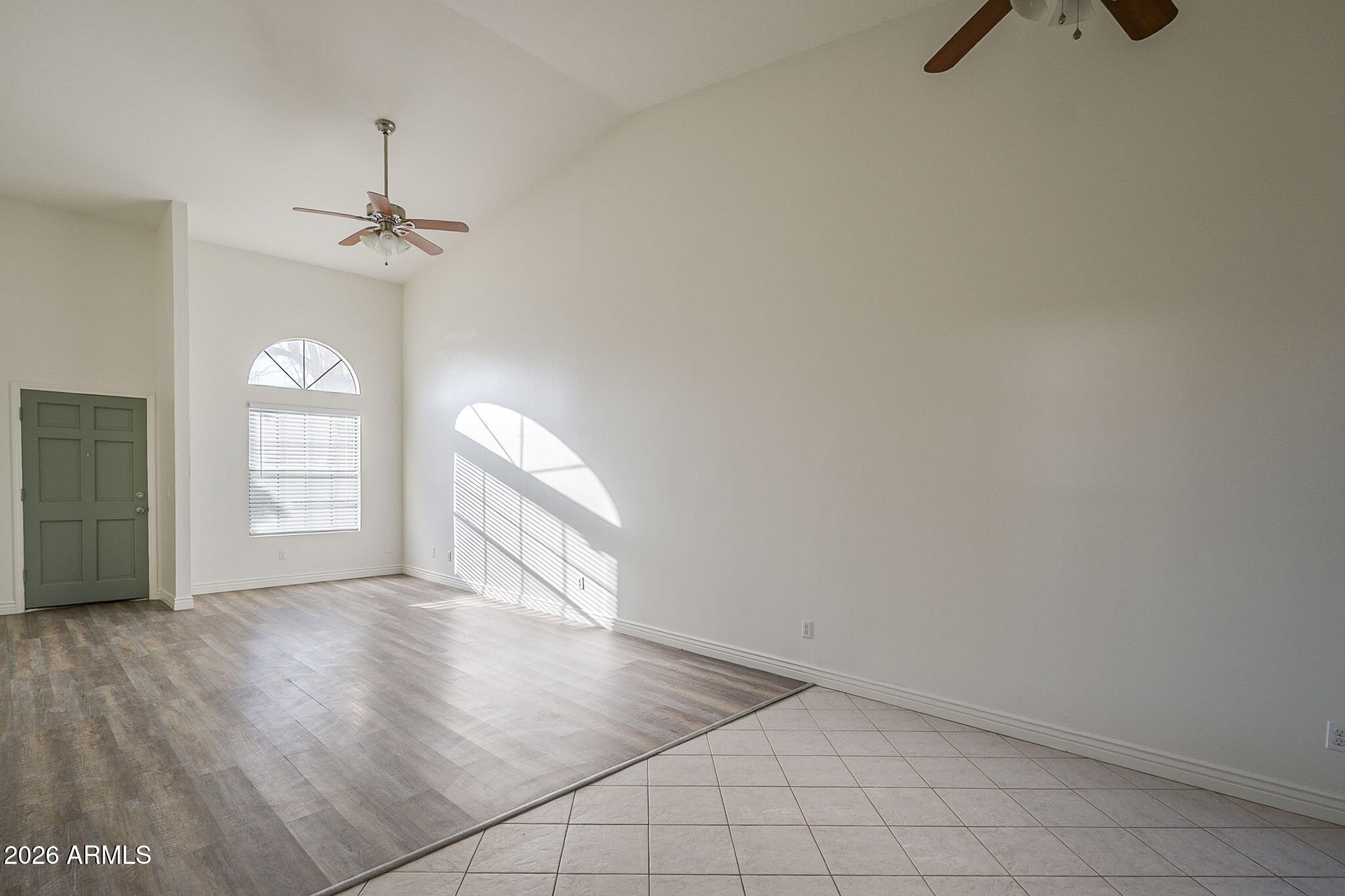 Empty living room with a high ceiling, wood-look flooring, and sunlight through tall windows.