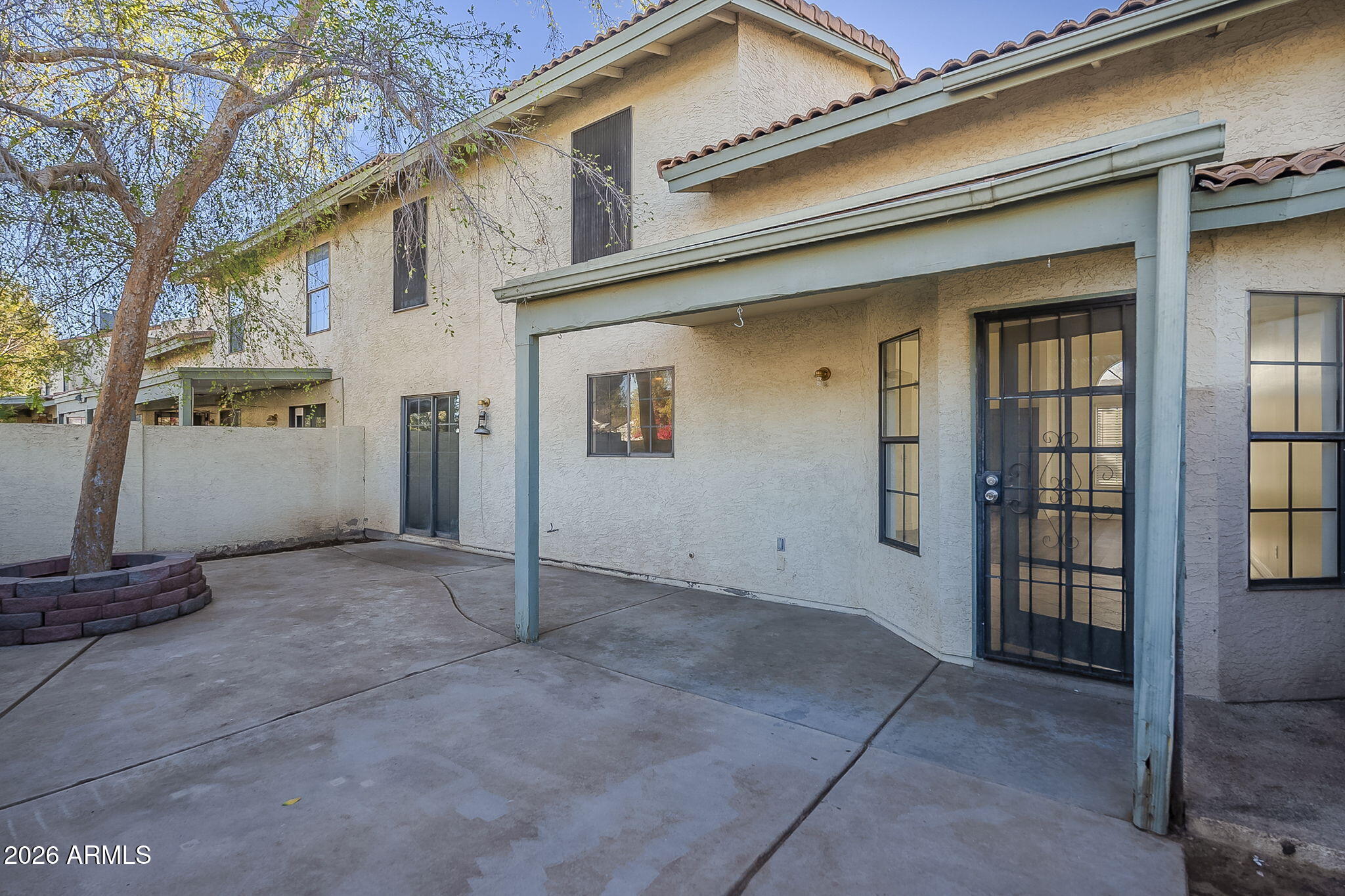 Home’s rear patio with a covered concrete area, security door, and mature tree in a raised planter.