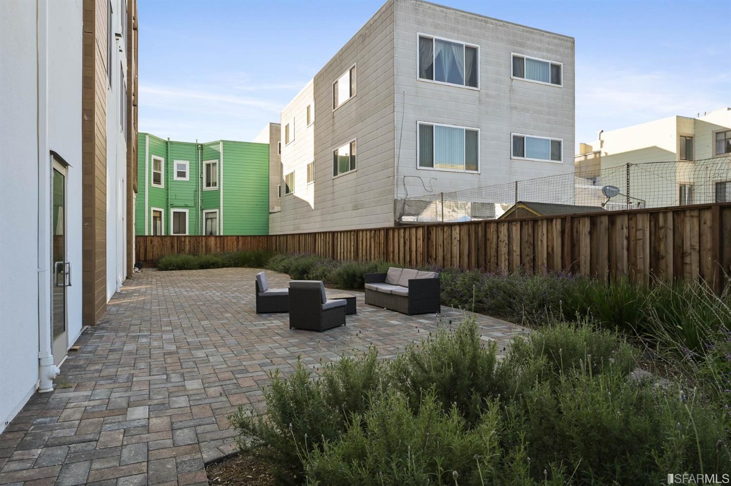 Residential courtyard with paved patio, landscaped beds, wooden fence, and wicker seating.