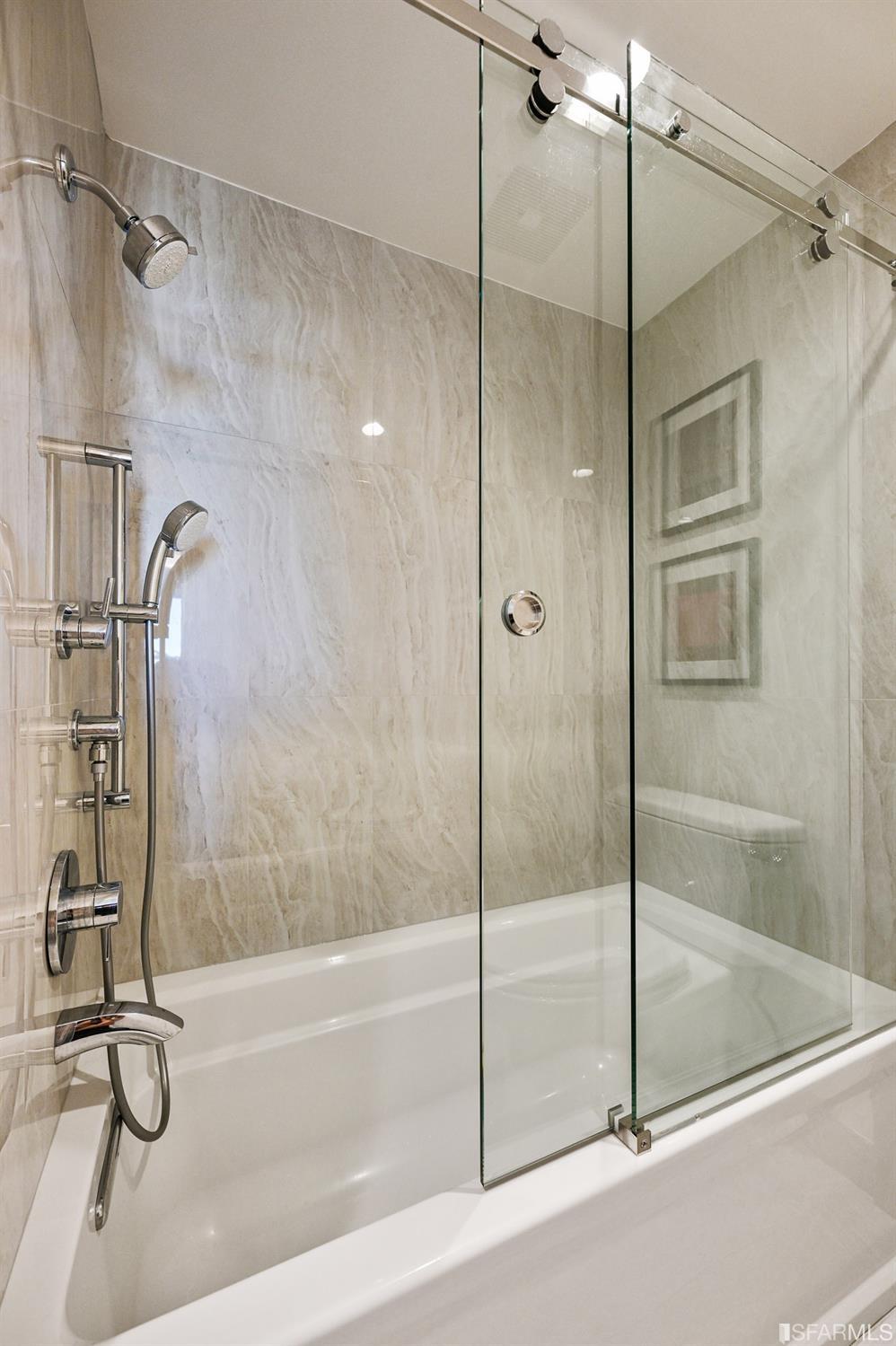 Bathtub with a clear glass shower enclosure, chrome fixtures, and beige marble-look tile.