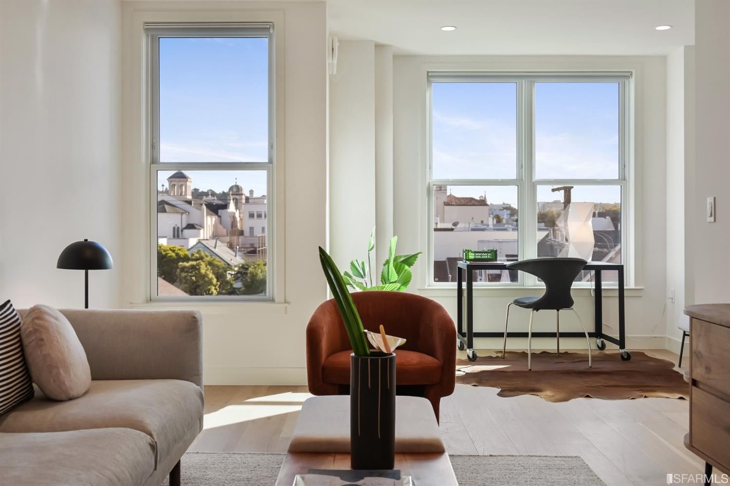 Bright living room with a gray sofa, rust-colored chair, and large windows overlooking city rooftops.