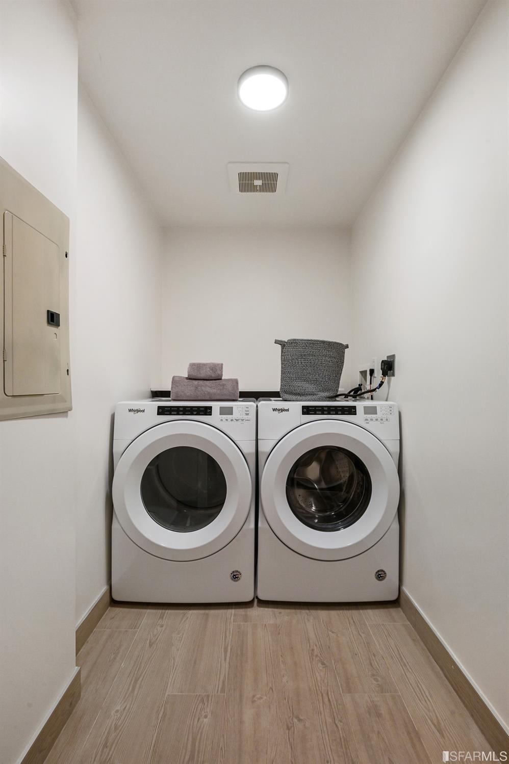 Laundry room with side-by-side front-loading washer and dryer, towels, basket, and wood-look tile flooring.