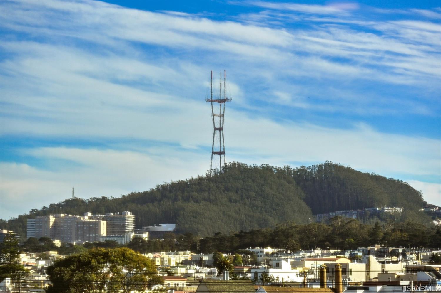 San Francisco rooftops with Sutro Tower on a wooded hill beneath a cloud-streaked sky.