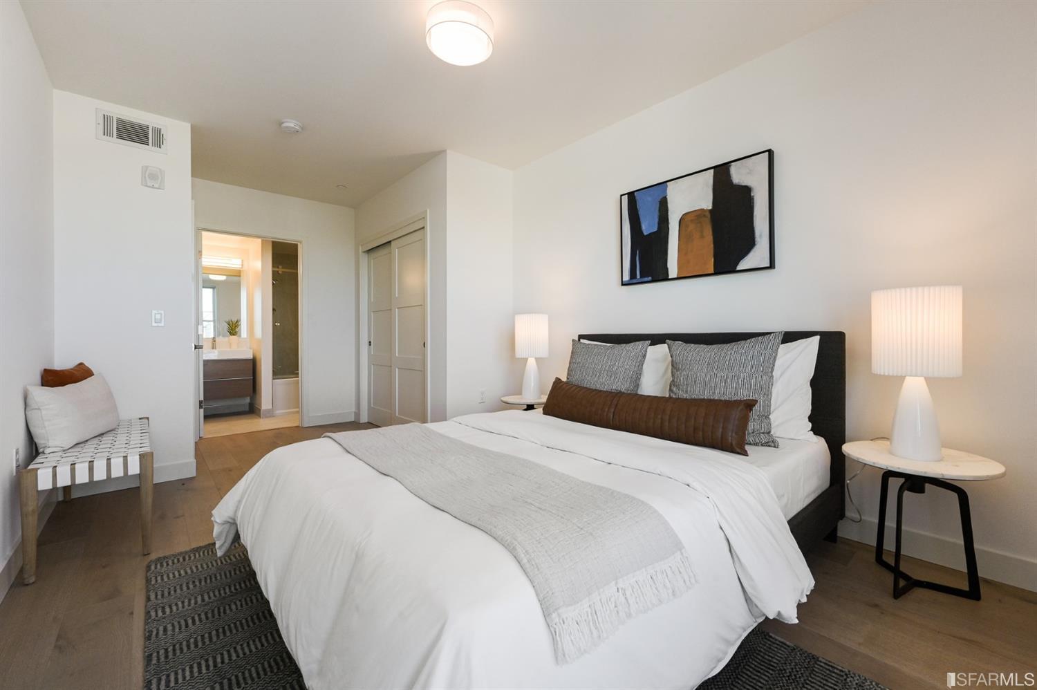 Staged bedroom with a white bed, matching bedside lamps, and an adjoining bathroom visible through the doorway.