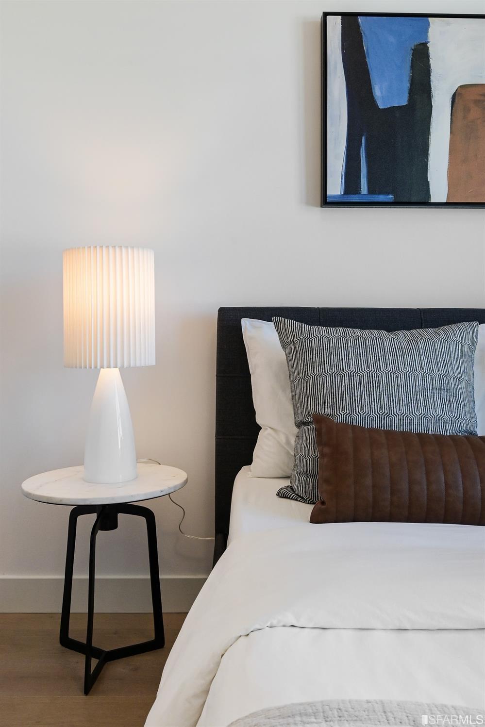 Staged bedroom with a white bed, patterned pillows, and a lit lamp on a marble side table.