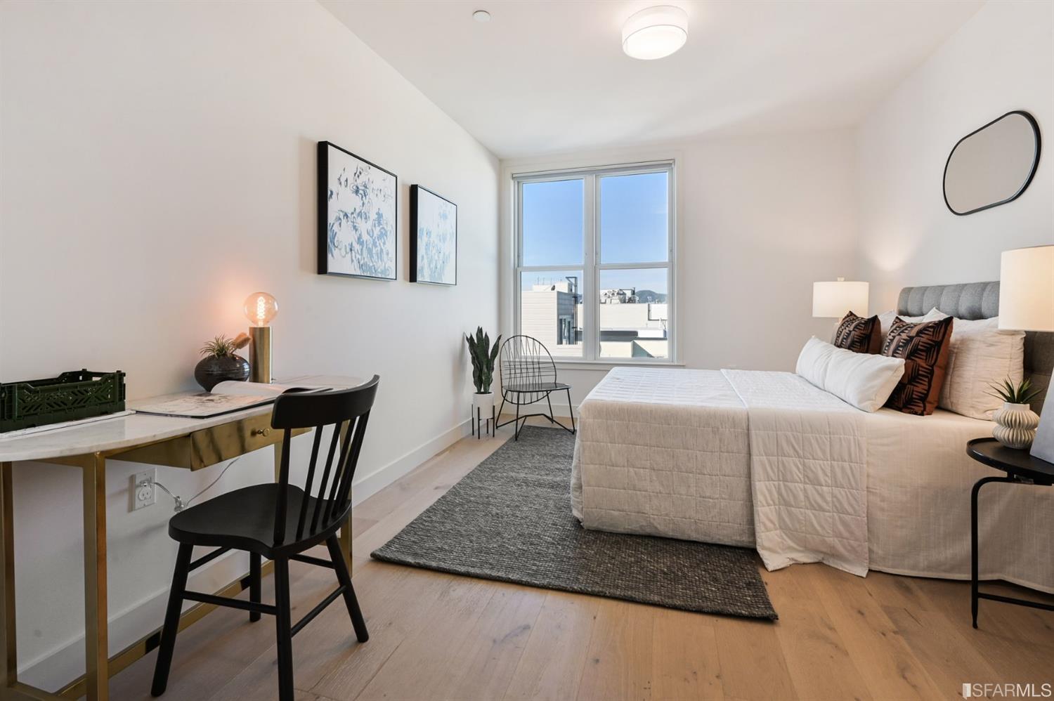 Staged bedroom with a large bed, desk, and tall window overlooking rooftops.