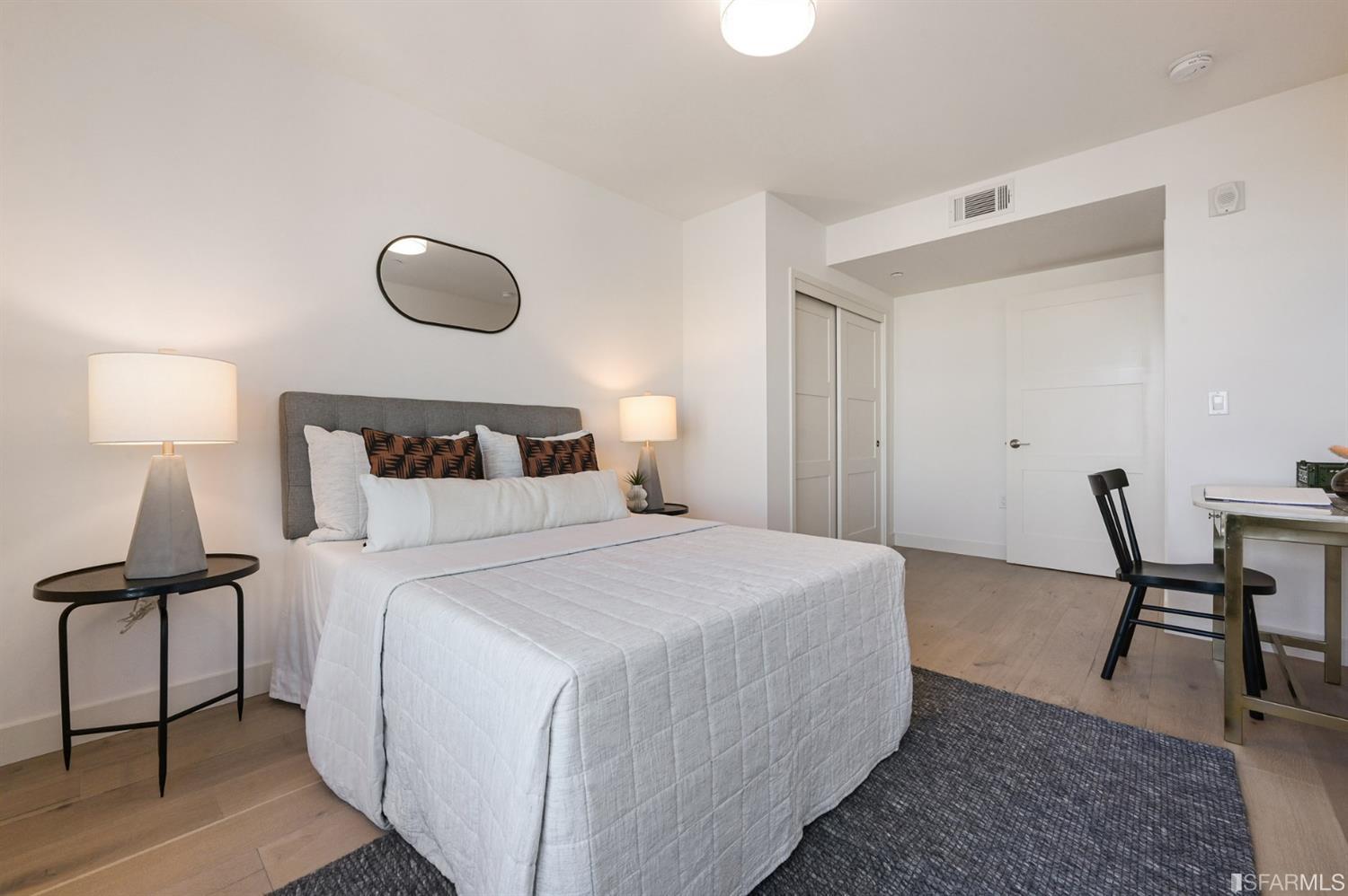 Staged bedroom with a white bed, gray upholstered headboard, bedside lamps, and dark area rug.