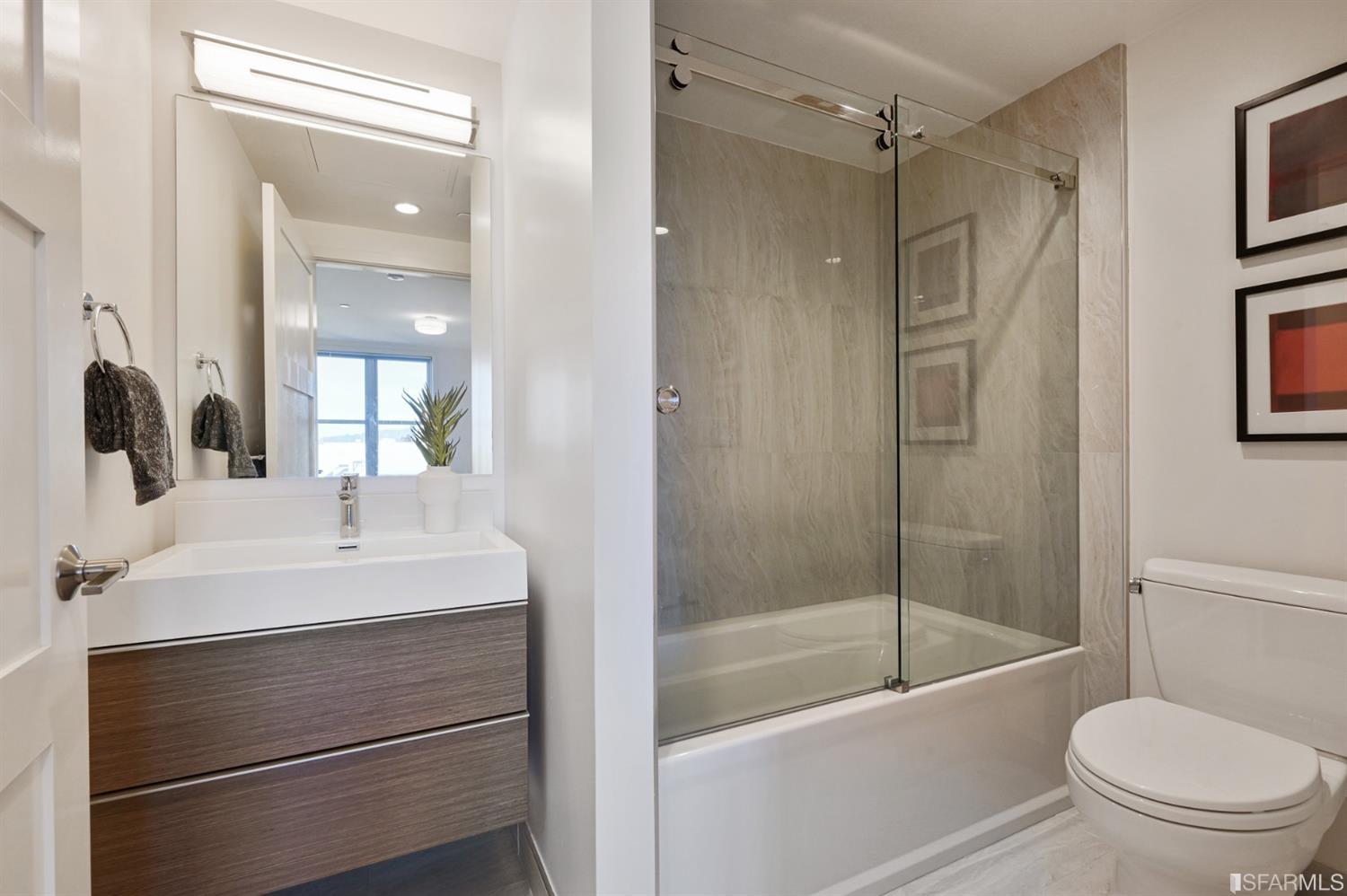 Bathroom with floating wood-tone vanity and a glass-enclosed tub with stone-look tile surround.