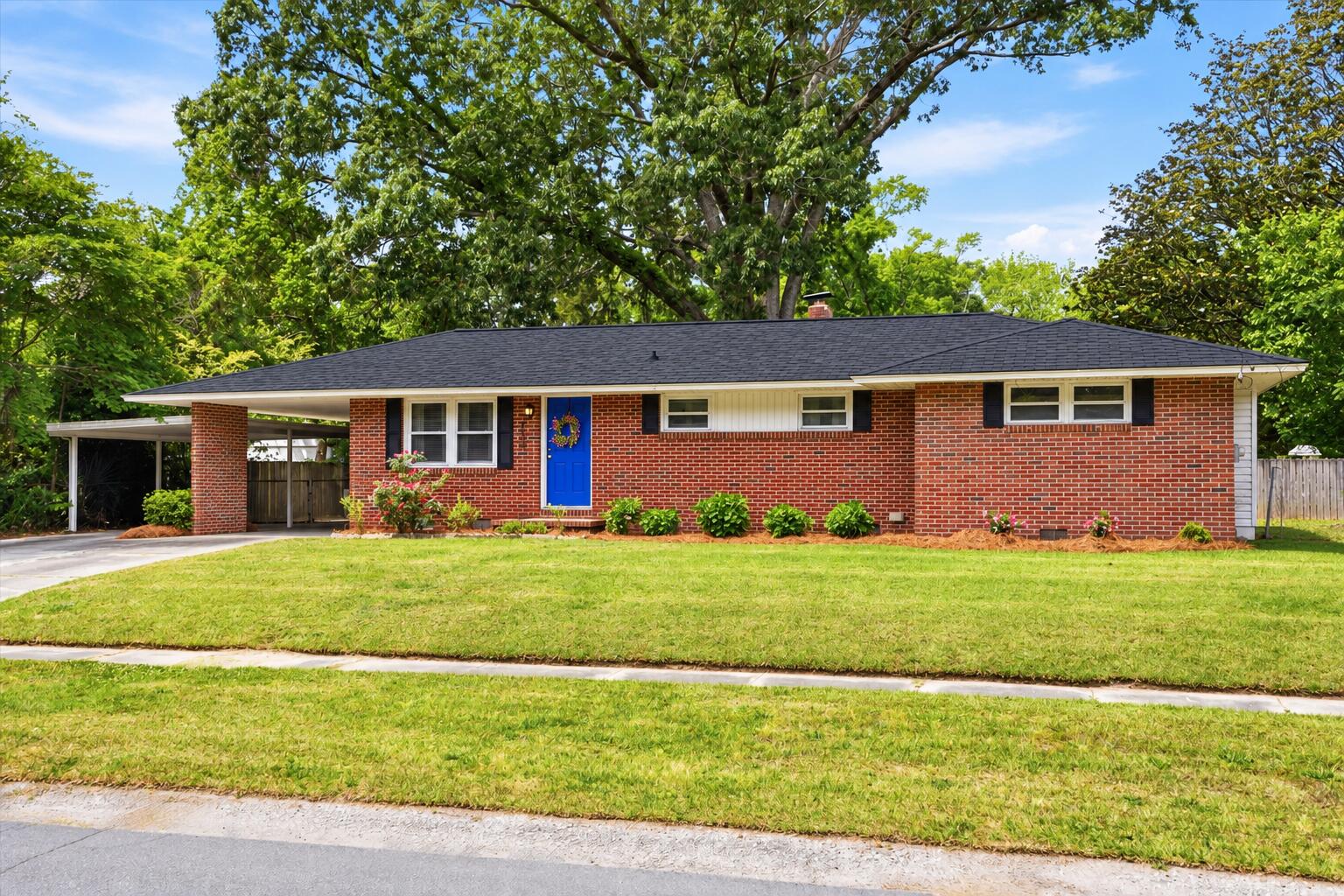 Red brick ranch house with a blue front door, covered carport, and mature trees.