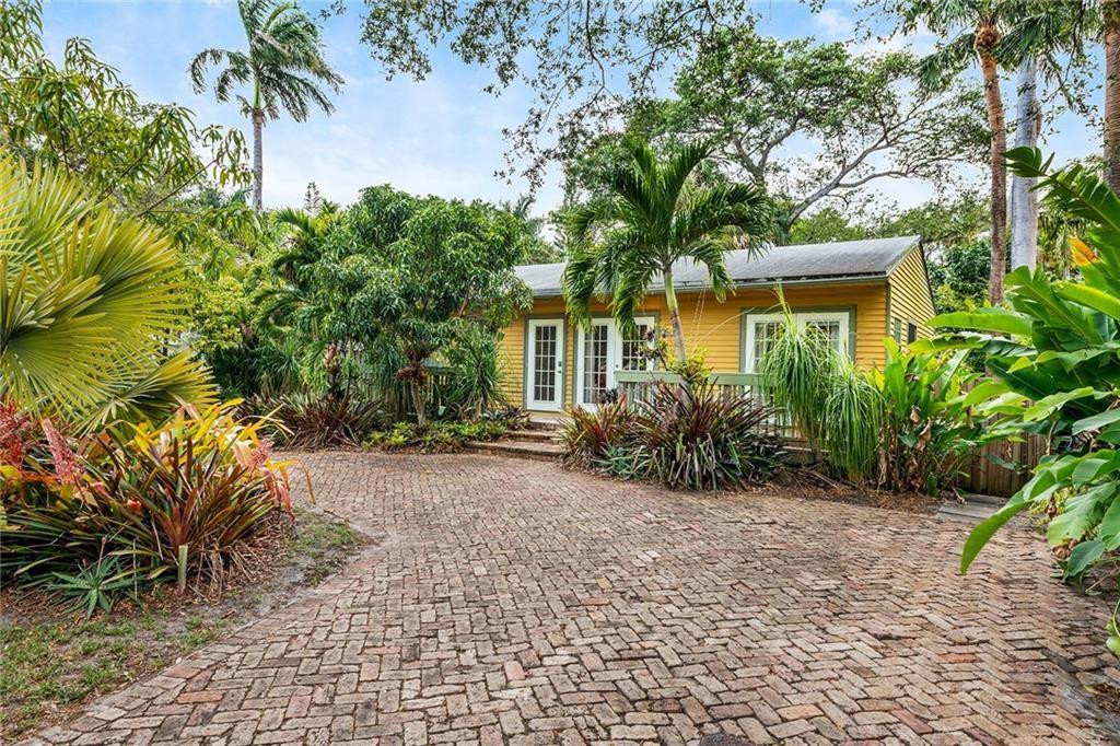 Yellow single-story home with white French doors, tropical landscaping, and a brick-paved driveway.