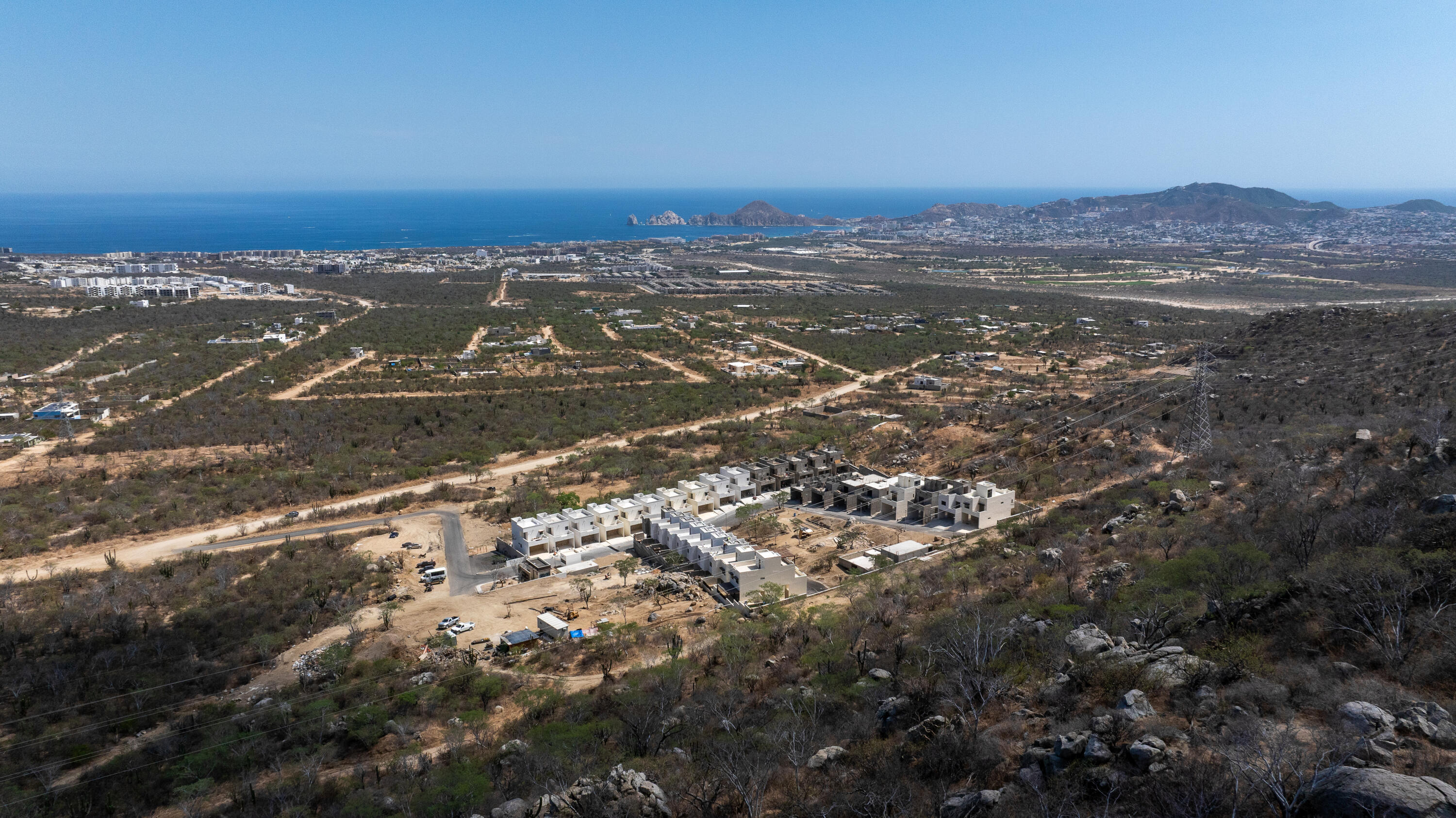 Aerial view of homes under construction on a rocky hillside, with the Pacific Ocean and coastal city beyond.