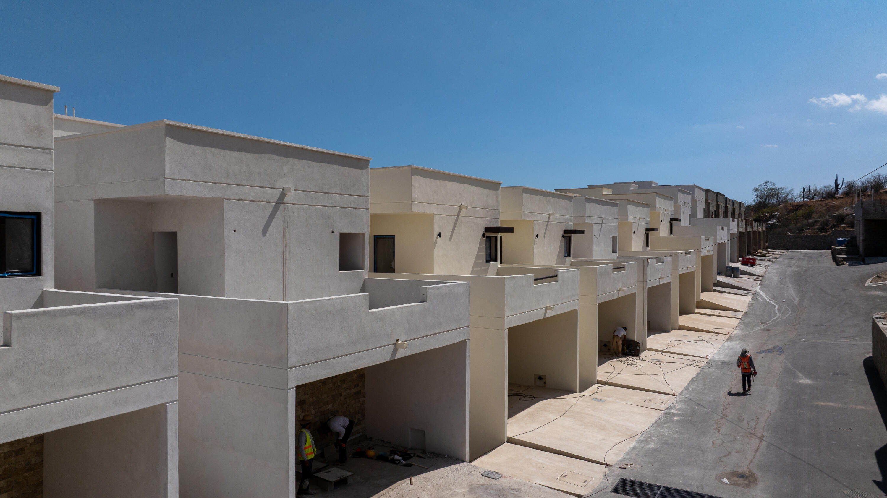 Row of modern homes under construction along a sloping street, with workers and building materials visible.