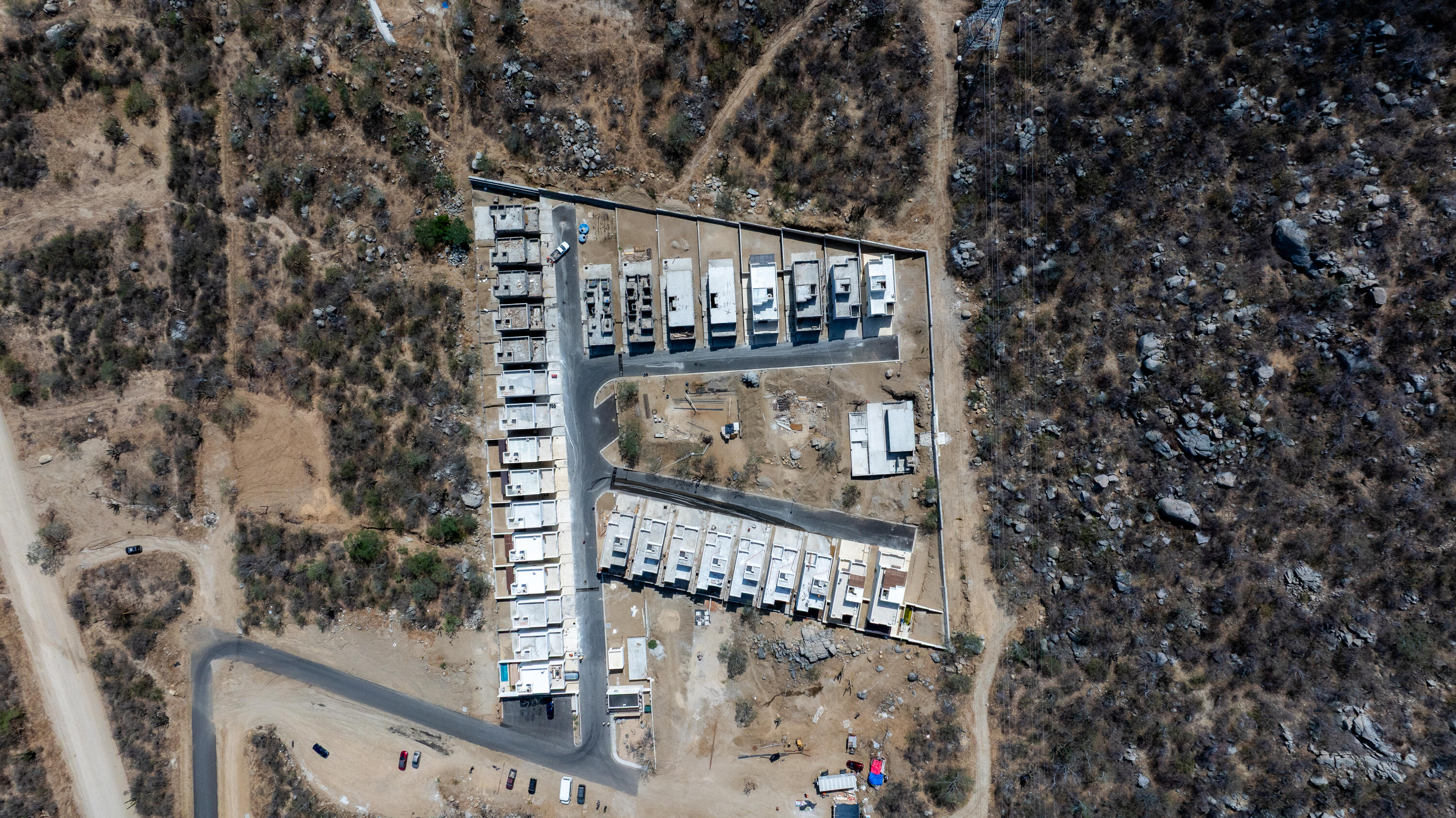 Aerial view of modern homes in rows amid rocky, dry terrain, with unfinished areas and construction materials.