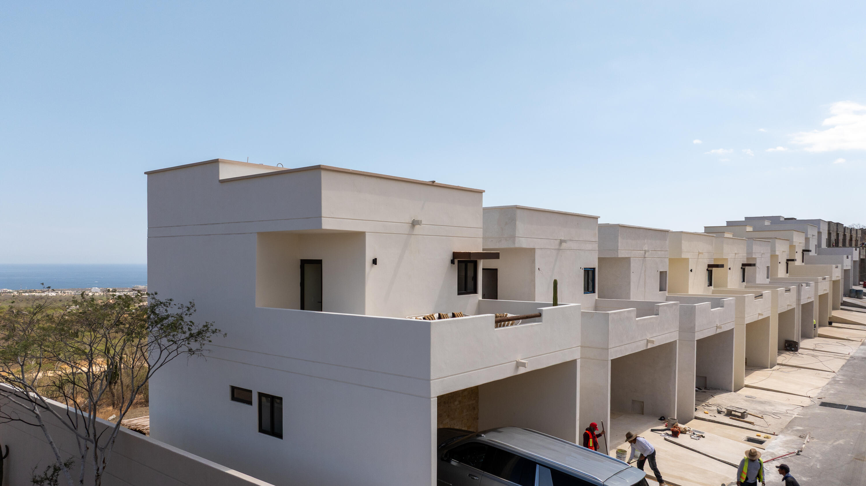 Modern white townhouses under construction, with workers in the foreground and ocean visible beyond.