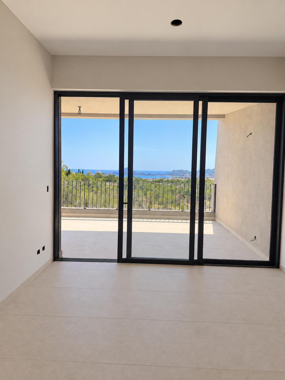 Empty tiled room with sliding glass doors opening to a balcony overlooking the sea and coastal hills.