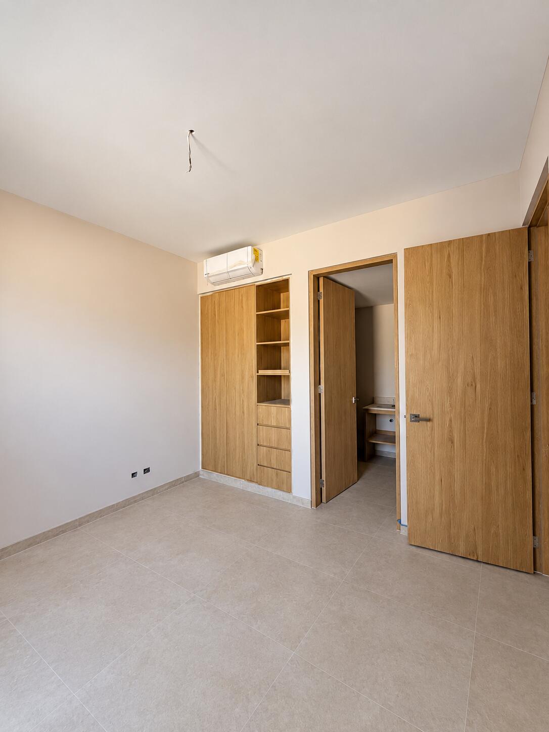Empty bedroom with built-in wooden wardrobe, beige tile floor, and wall-mounted air conditioner.