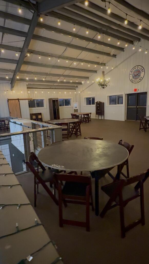 Spacious venue with exposed beams, string lights, round tables, and folding chairs.