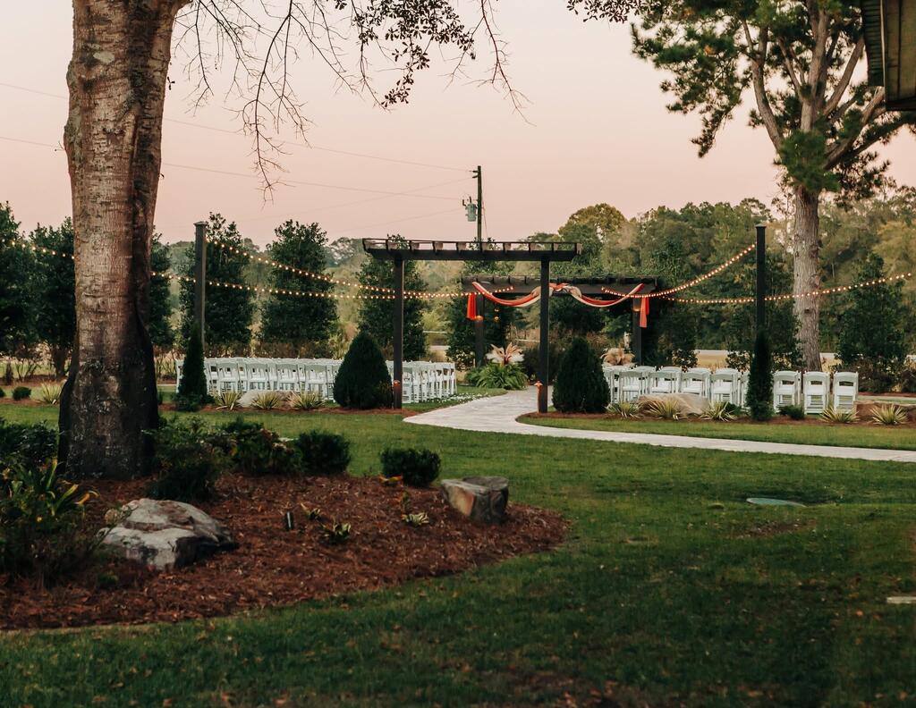 Wedding ceremony setup with a draped wooden arbor, string lights, white chairs, and a curved garden walkway.