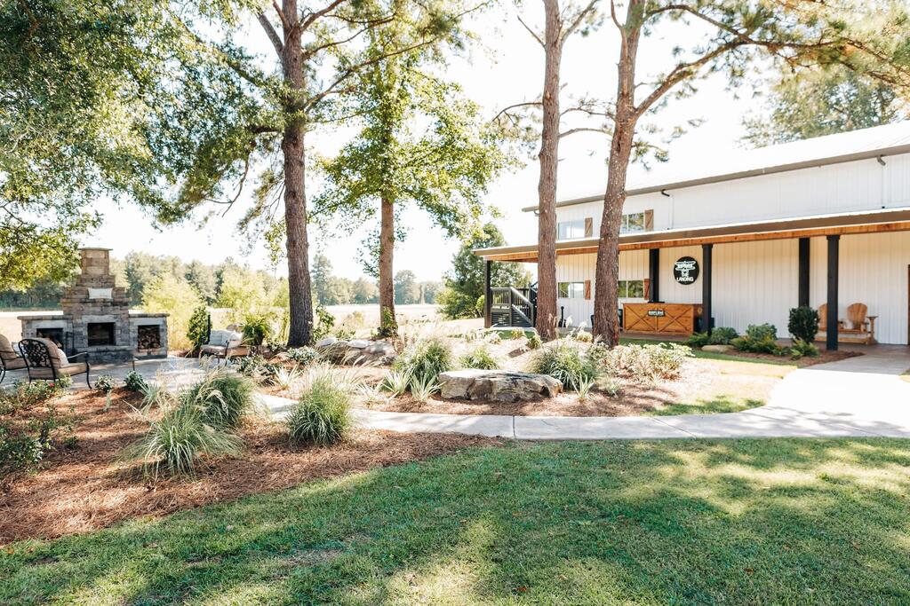 White property building with a covered porch, landscaped lawn, and stone outdoor fireplace beneath mature trees.
