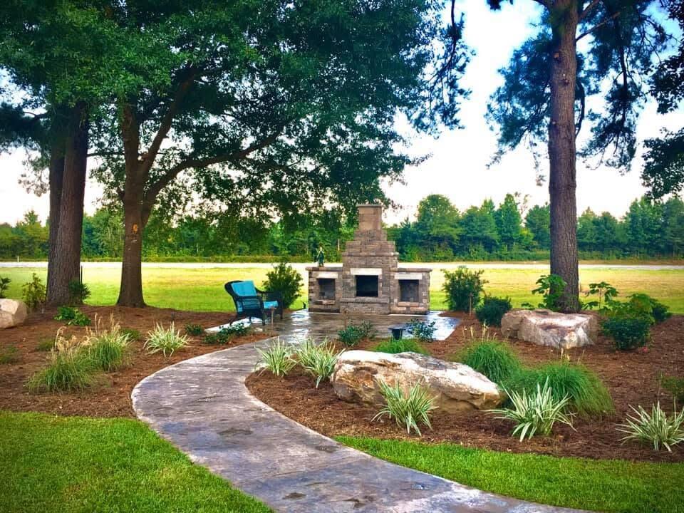 Landscaped patio with a stone fireplace, blue chair, and curved walkway beneath mature trees.