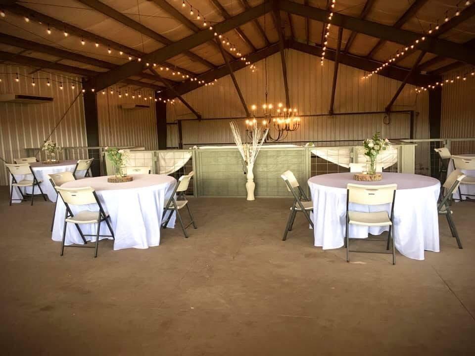 Event space with white-clothed round tables, folding chairs, and string lights beneath exposed roof trusses.