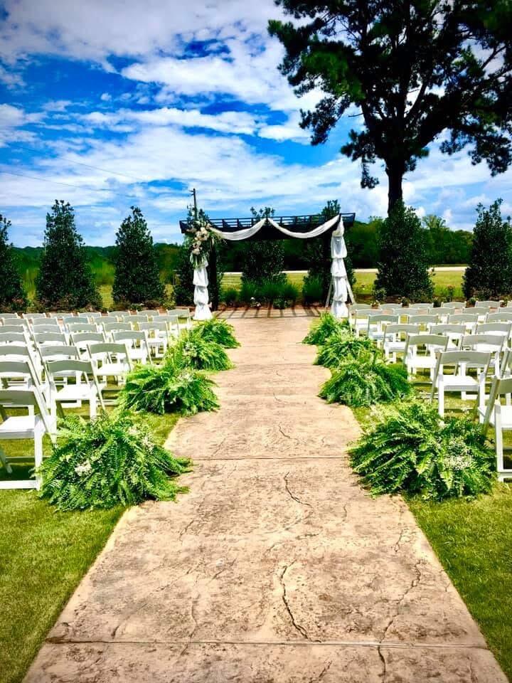 Outdoor ceremony aisle lined with white chairs and ferns, leading to a draped arch.