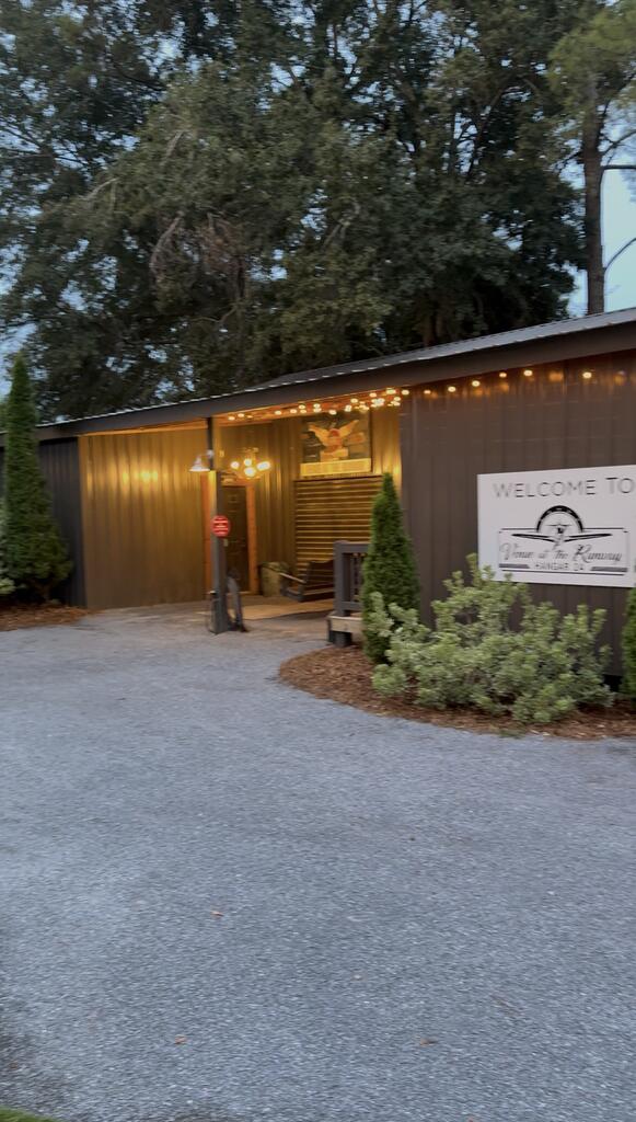 Dark wood building entrance with warm string lights, gravel drive, and a welcome sign.