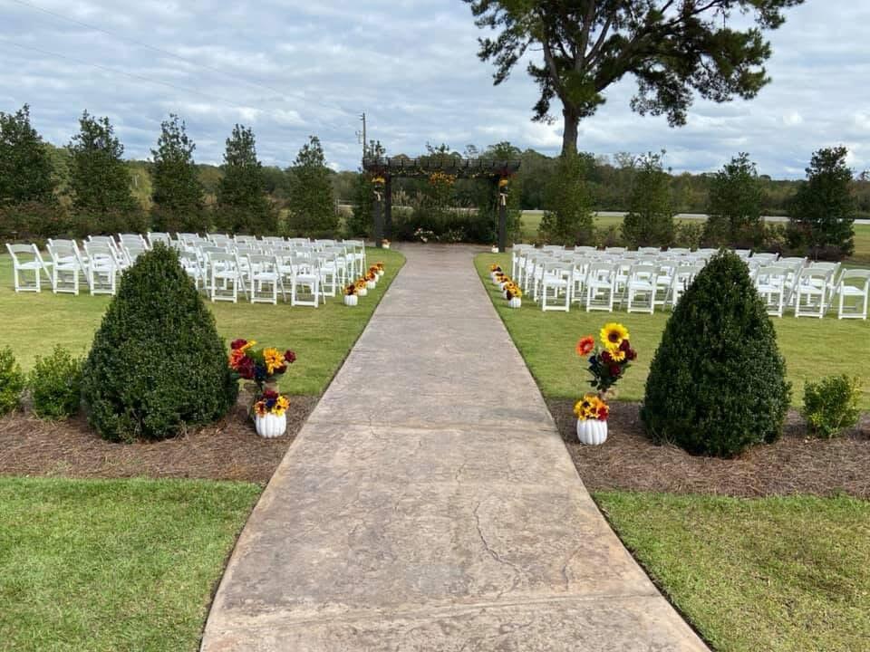 Outdoor ceremony space with white chairs, a concrete aisle, and a wooden arbor.