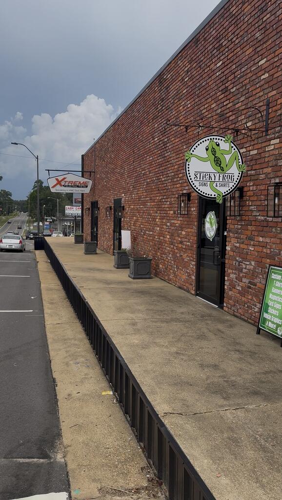 Brick storefront with a Sticky Frog Signs & Shirts sign above the entrance and a raised sidewalk.