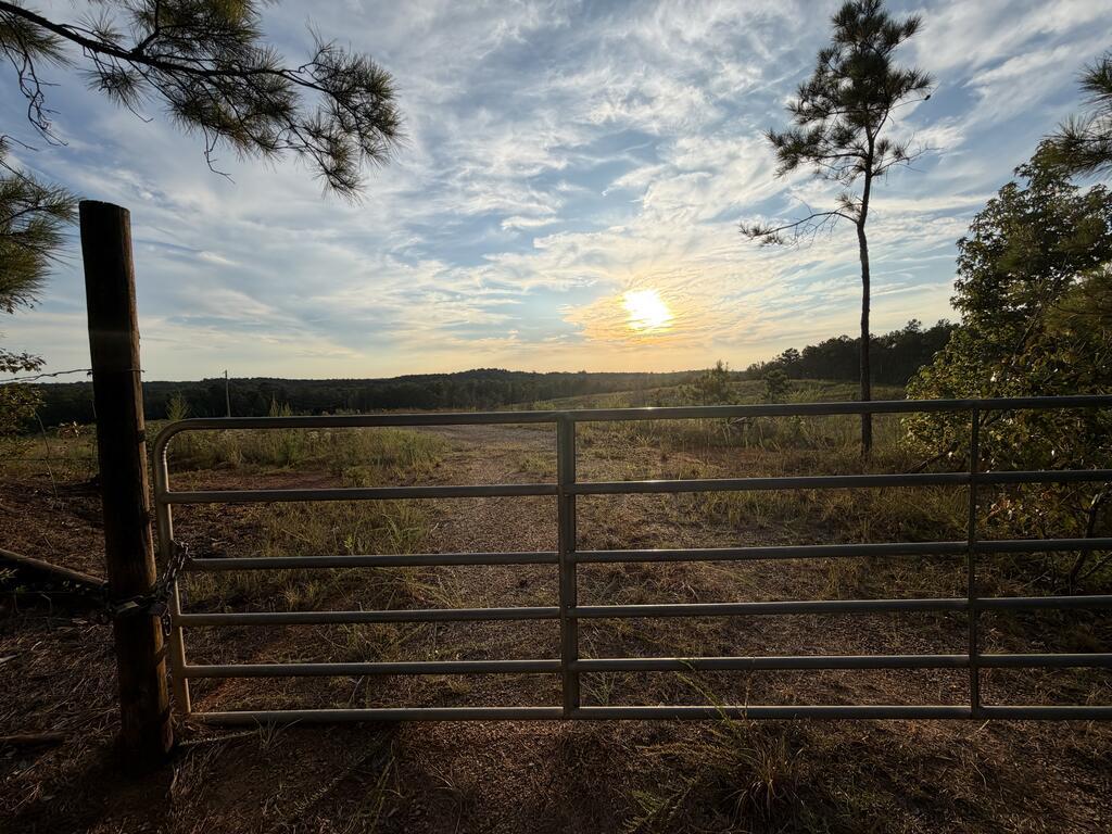 Rural land entrance with a metal gate, gravel track, open fields, and wooded hills at sunset.