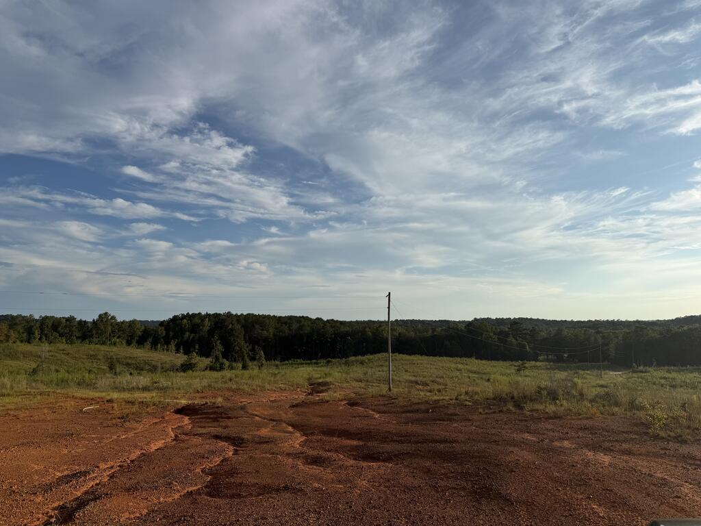 Vacant rural land with red dirt, rolling grass, distant trees, and utility poles.