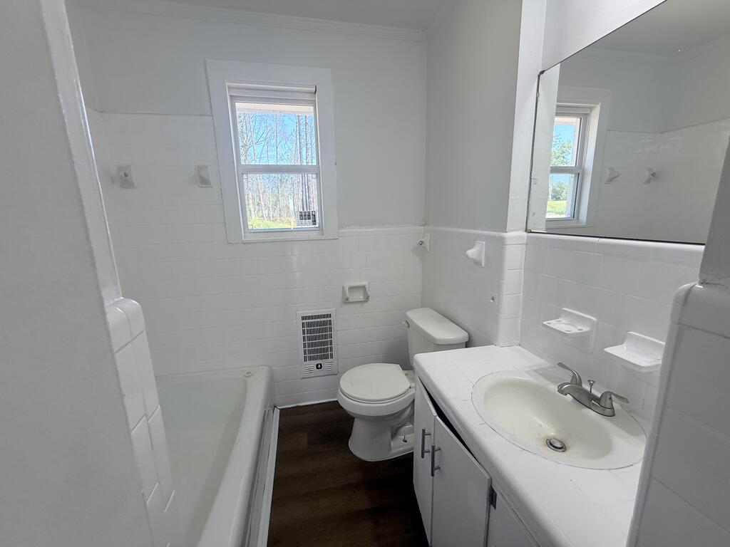 Vacant bathroom with a bathtub, toilet, white tiled walls, and a vanity beneath a large mirror.