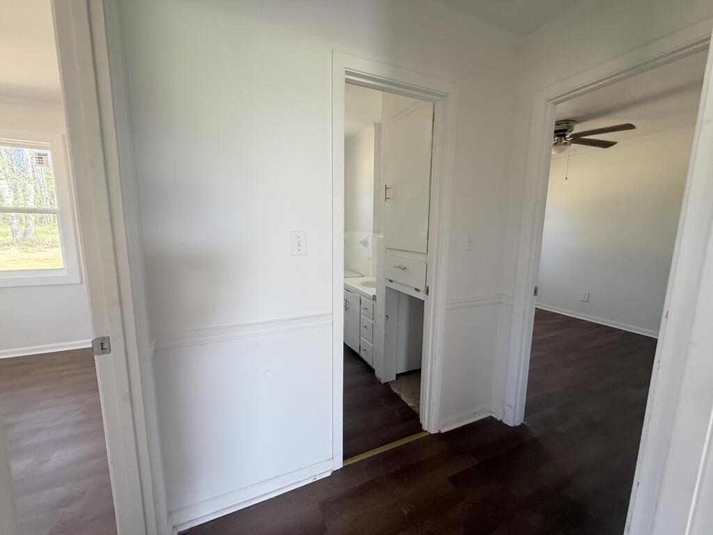 Hallway with dark flooring connects a bathroom and an empty room with a ceiling fan.