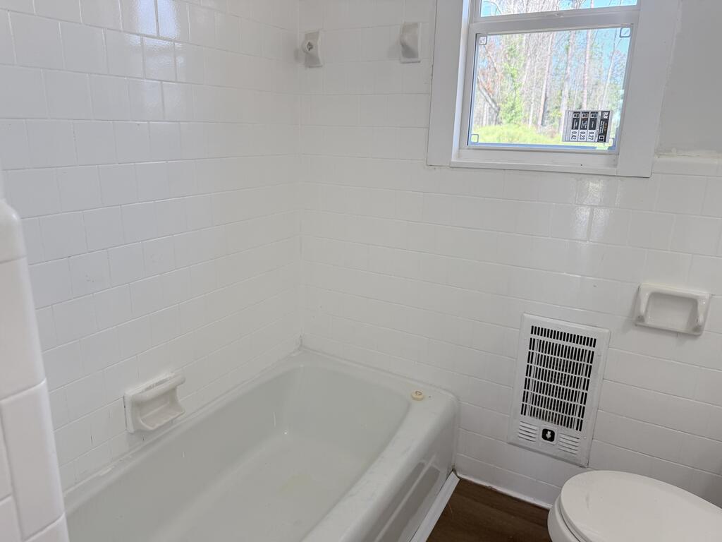 White tiled bathroom with a bathtub, toilet, and window overlooking trees.