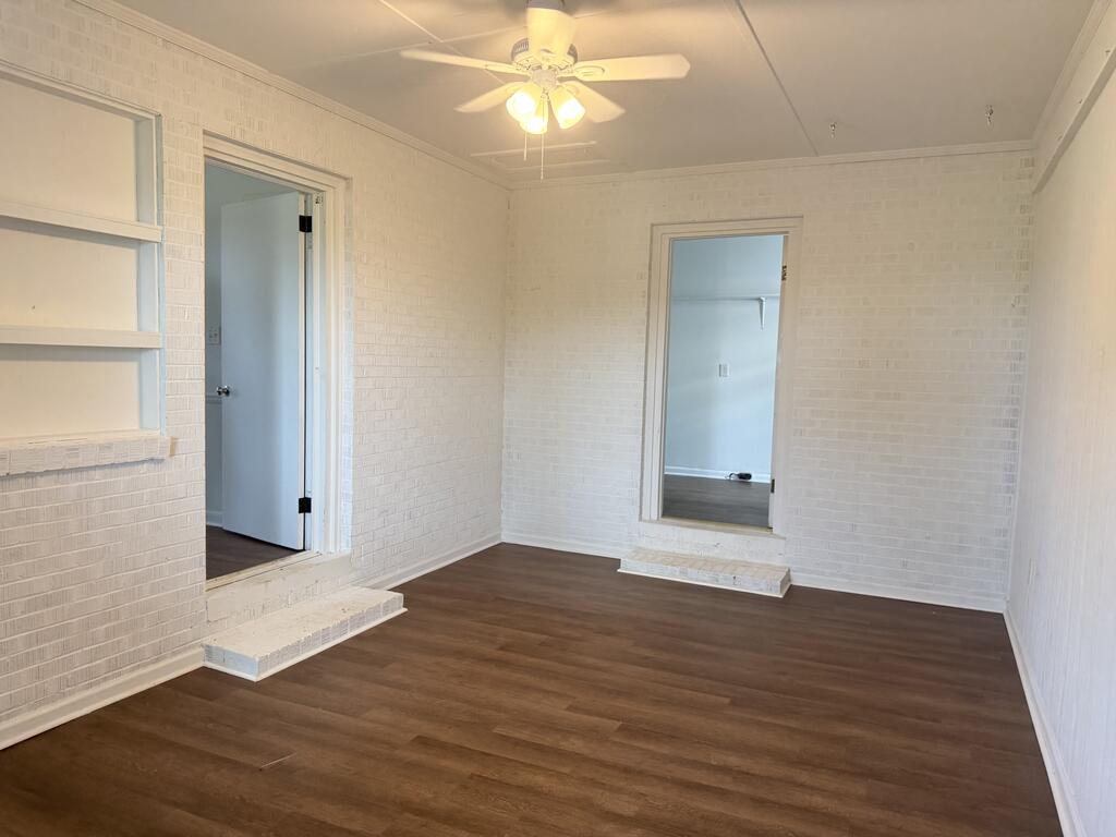 Empty room with white brick walls, dark flooring, built-in shelves, and a ceiling fan.