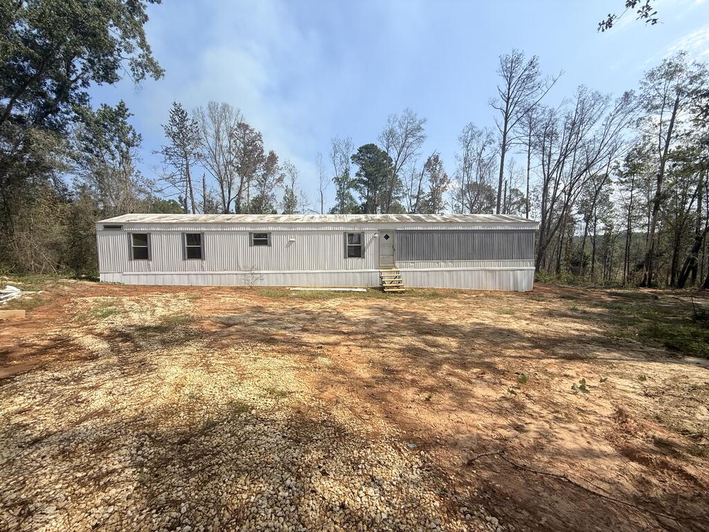 Long metal-sided manufactured home in a gravel clearing surrounded by trees.