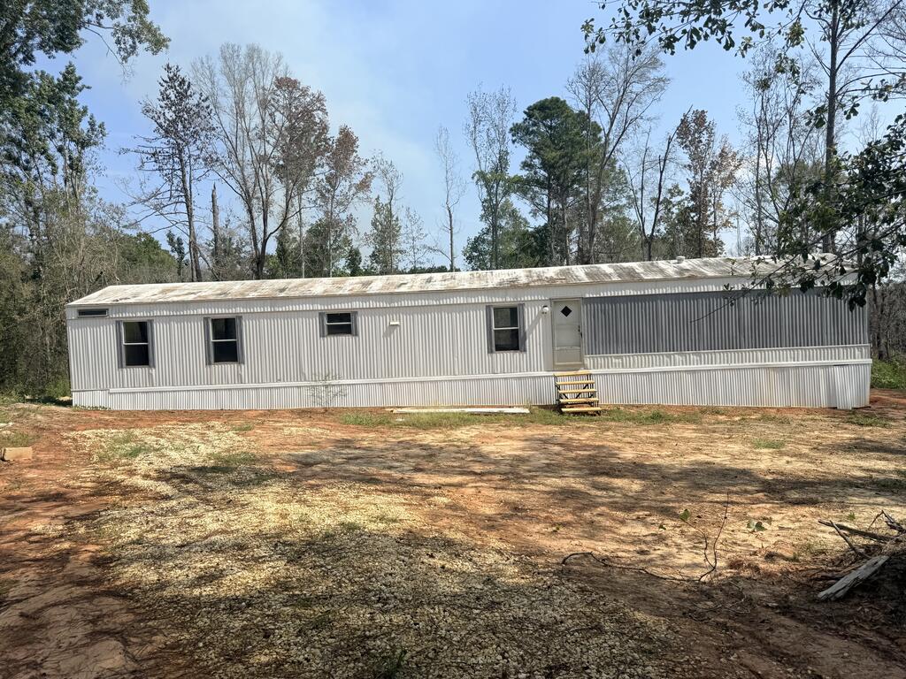 Long gray metal-sided manufactured home with front windows and entrance steps in a wooded clearing.