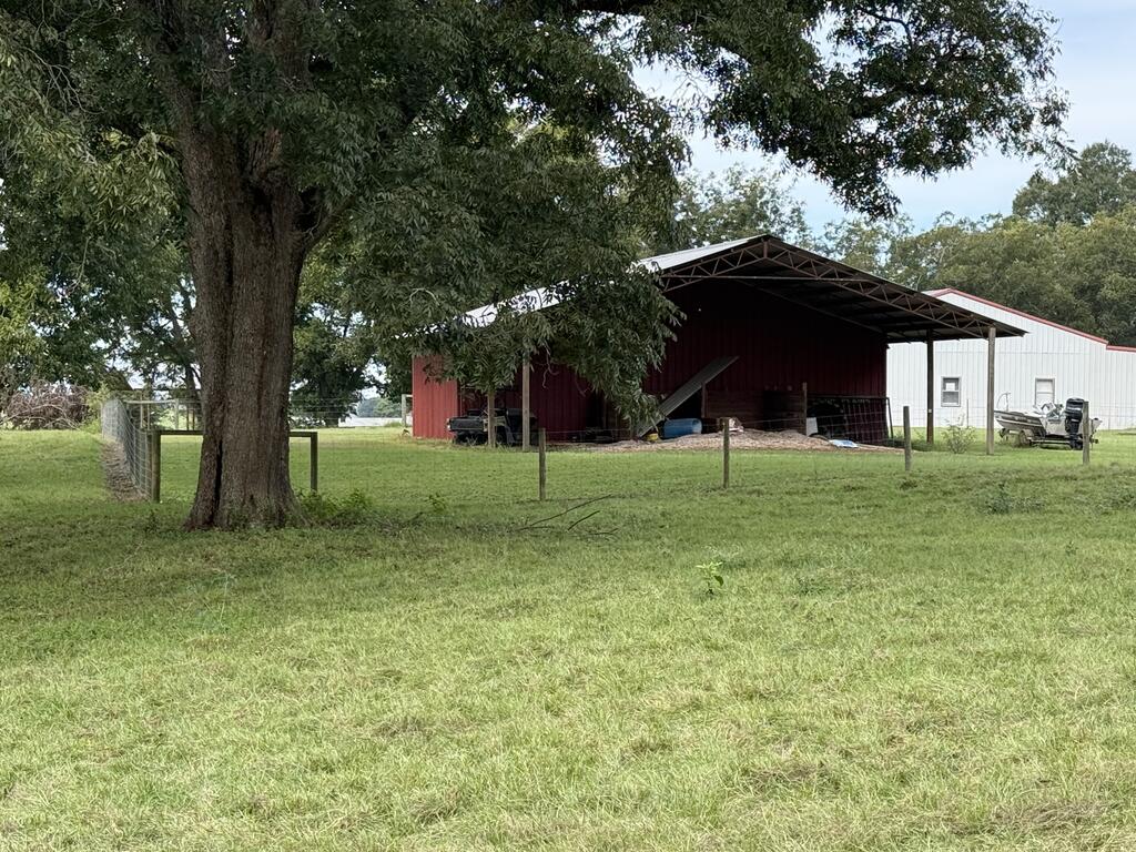 Red open-sided barn and white outbuilding behind a fence, with a large shade tree in front.