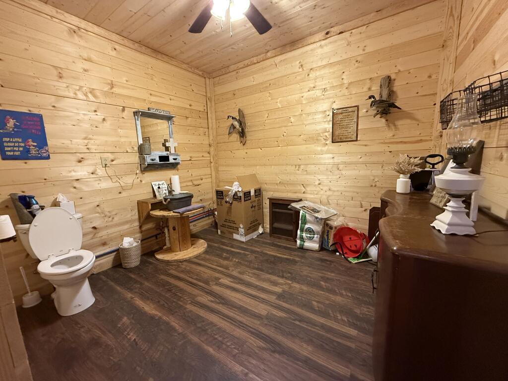 Bathroom with pine plank walls, a toilet, and assorted items on dark wood-look flooring.