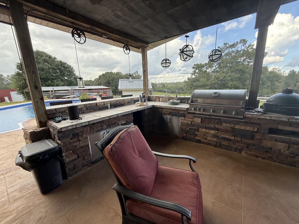 Covered outdoor kitchen with stone counters and built-in grills beside a swimming pool.