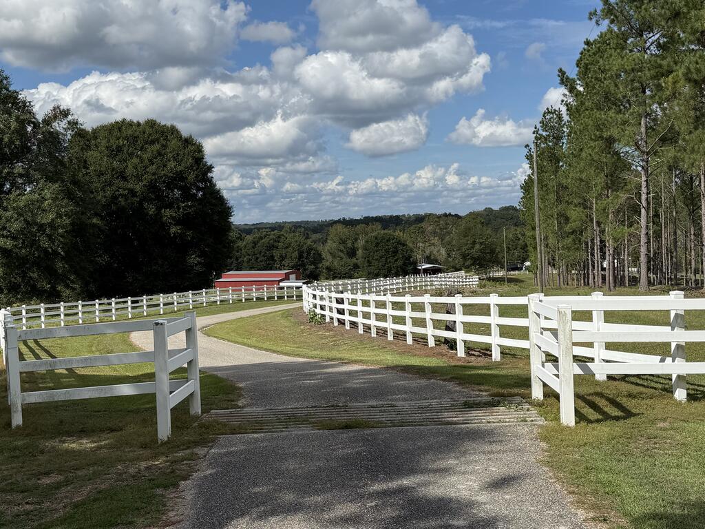 Rural driveway entrance bordered by white fencing, with a red barn and woods beyond.
