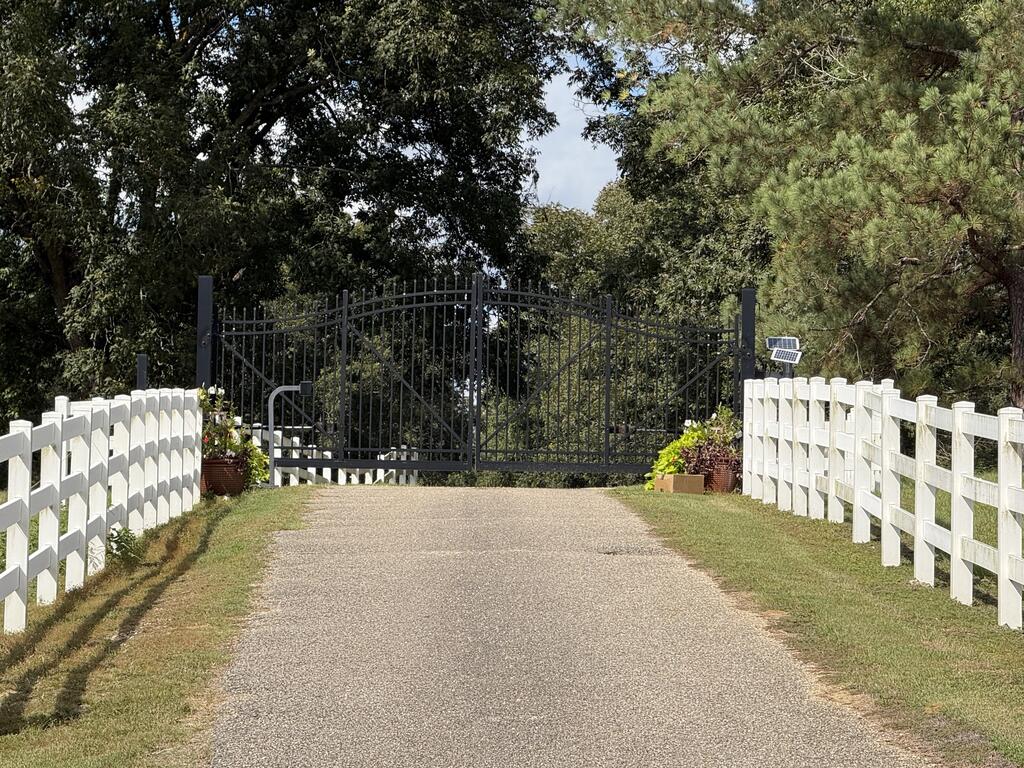 Gated gravel driveway bordered by white fences and mature trees.