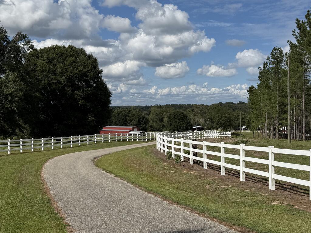 Rural property driveway curving between white rail fences toward a red-roofed outbuilding.